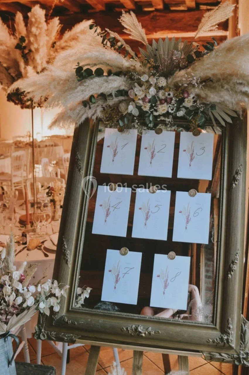 Plan de table de mariage affiché dans un cadre doré, décoré de fleurs séchées et de pampas, dans une salle élégante.