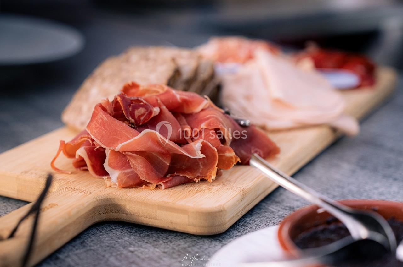 Planche en bois garnie de tranches de jambon cru, pain et autres charcuteries, posée sur une table grise.
