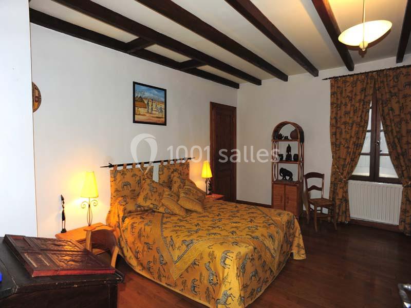 Chambre avec lit double, couvre-lit jaune à motifs, poutres apparentes, mobilier en bois et lumière tamisée.