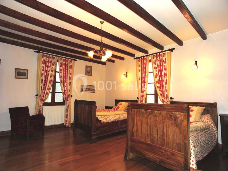 Chambre rustique avec poutres apparentes, lits en bois, parquet et rideaux rouges à motifs, éclairée par des appliques…