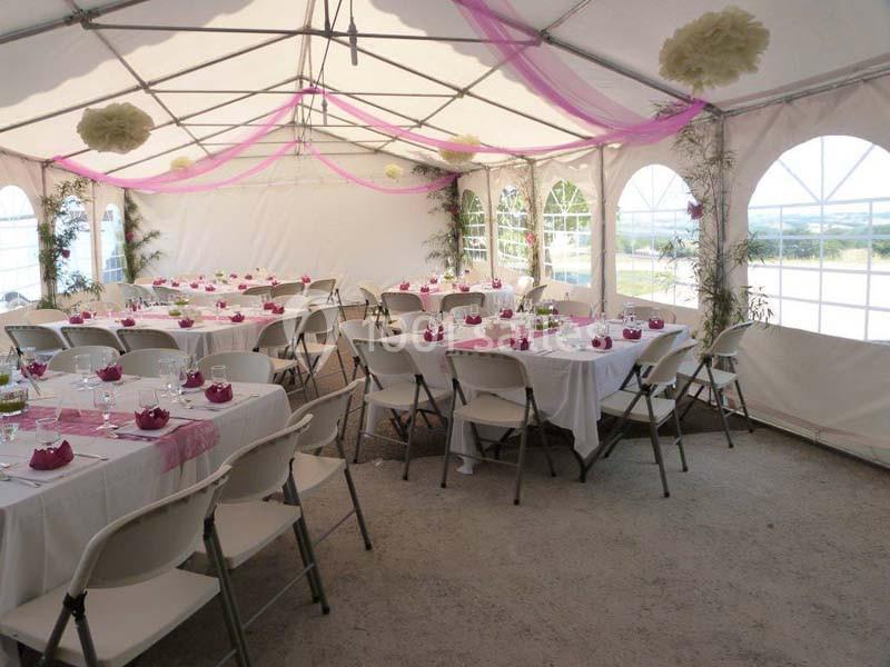 Salle de réception sous une tente blanche avec tables dressées, décorations roses et chaises pliantes.