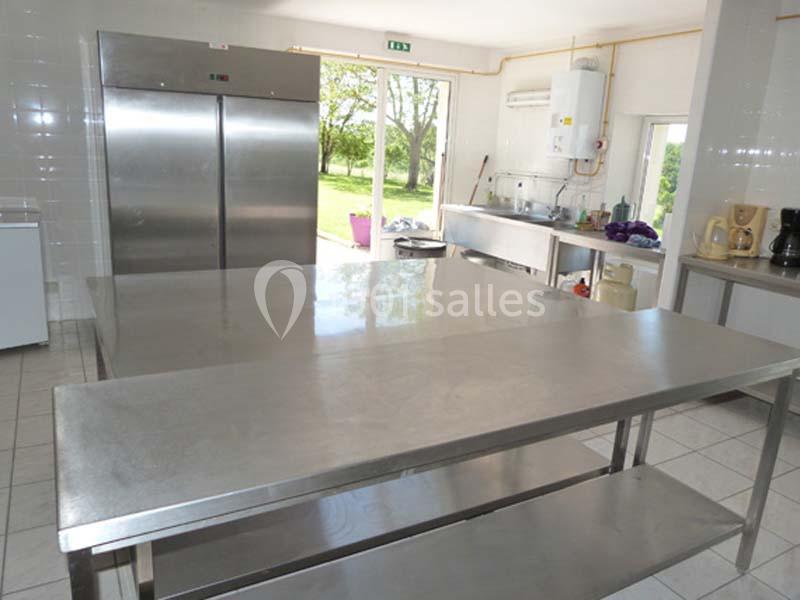 Cuisine professionnelle avec plans de travail en inox, réfrigérateurs, évier et vue sur un jardin par une porte vitrée.