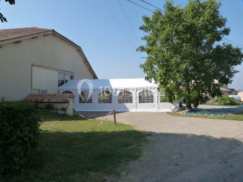 Tente blanche installée à côté d'un bâtiment, entourée d'un arbre, d'herbe et d'une allée en gravier.