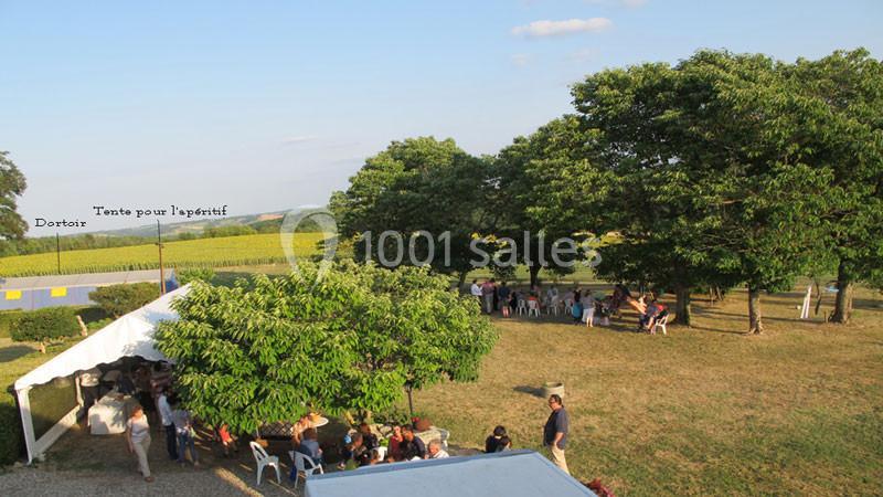 Groupe de personnes rassemblées sous des arbres et une tente dans un espace extérieur ensoleillé près de champs.