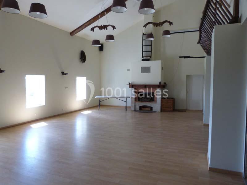 Salle lumineuse avec parquet, cheminée, poutres apparentes et mezzanine, éclairée par des fenêtres et des suspensions.