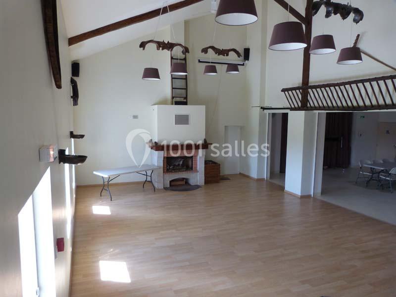 Salle lumineuse avec parquet clair, cheminée en pierre, poutres apparentes et suspensions modernes au plafond.