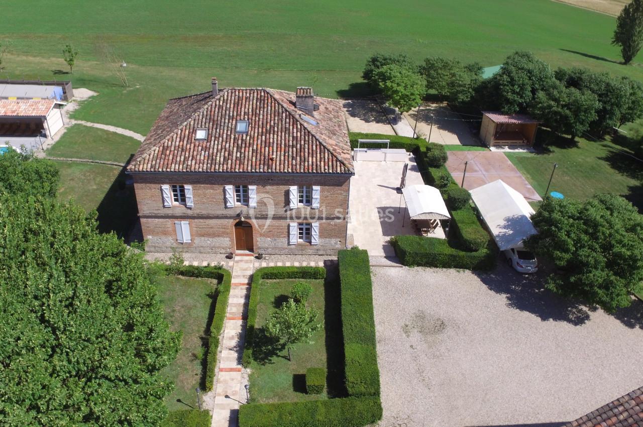 Vue aérienne d'une maison en pierre avec toit en tuiles, entourée de jardins, d'arbres et d'une cour gravillonnée.