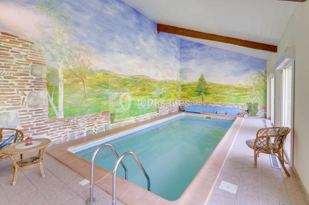 Piscine intérieure entourée de murs peints représentant un paysage champêtre, avec chaises en osier et lumière naturelle.