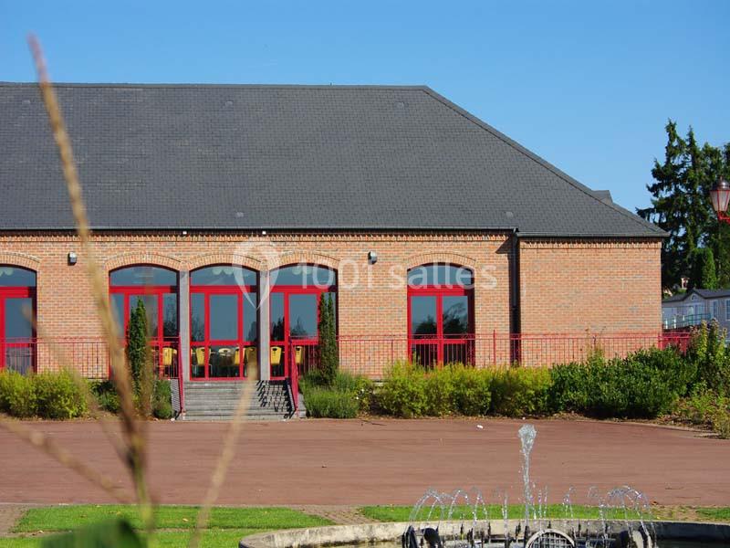 Bâtiment en briques rouges avec des portes et fenêtres rouges, entouré de végétation et d'une fontaine au premier plan.