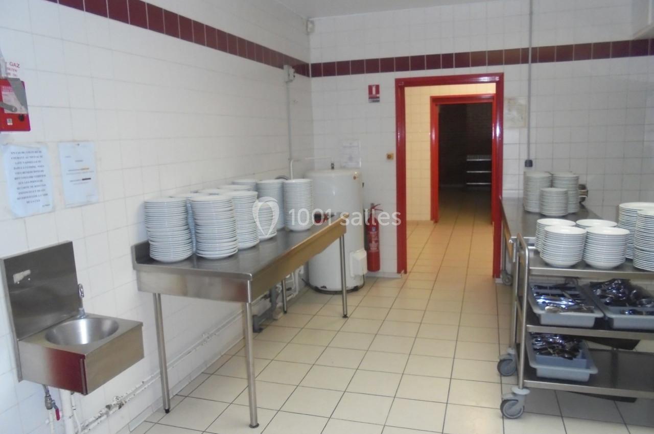 Cuisine professionnelle avec piles d'assiettes blanches sur des tables en inox et chariots contenant des couverts.