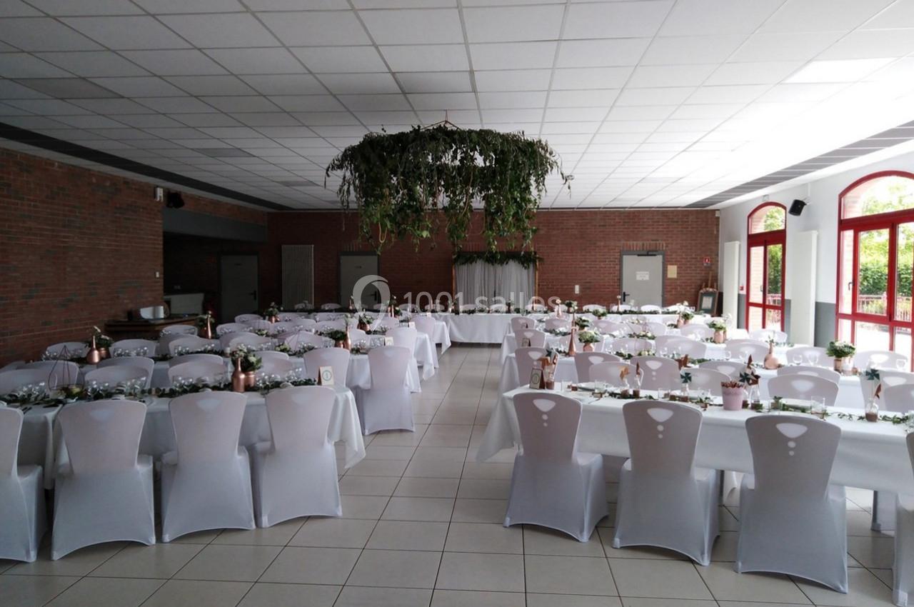 Salle de réception décorée avec des tables alignées, nappes blanches, chaises houssées et centre de table végétal suspendu.
