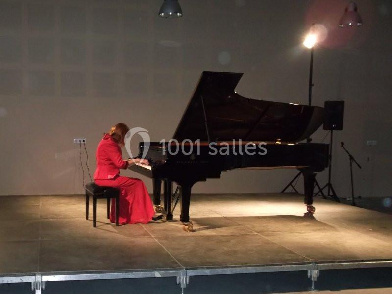 Une femme en robe rouge joue du piano à queue sur une scène éclairée dans une salle sombre.