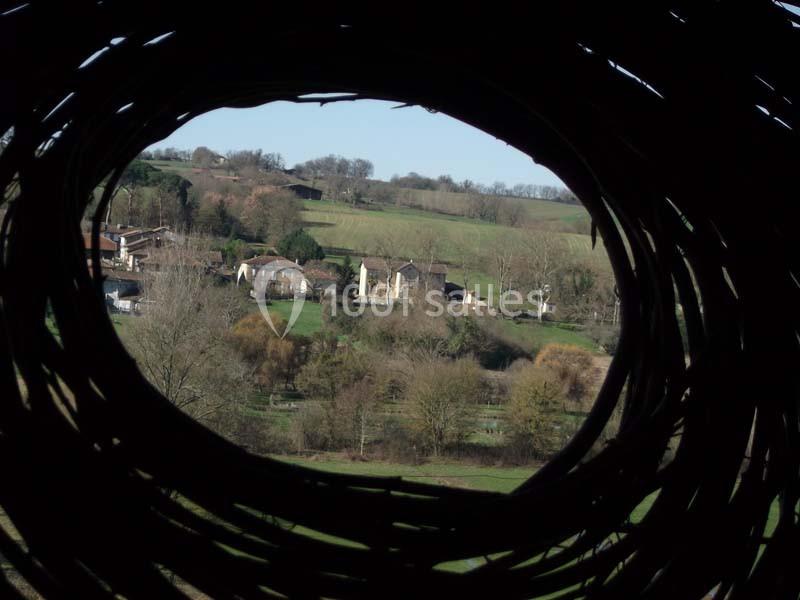 Vue d'un village rural et de collines verdoyantes à travers une ouverture circulaire en branches tressées.