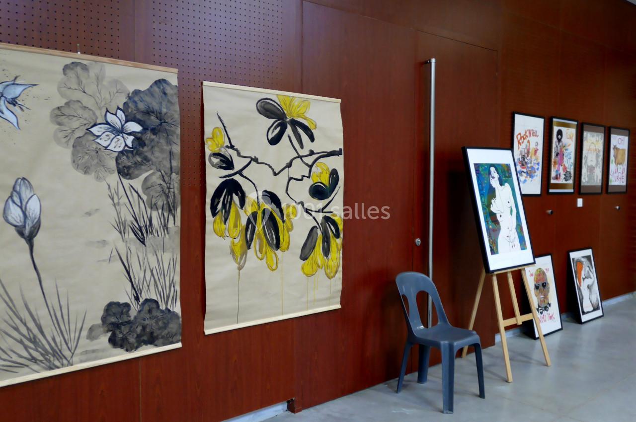 Exposition de peintures et dessins accrochés sur un mur en bois, avec une chaise et un chevalet au premier plan.