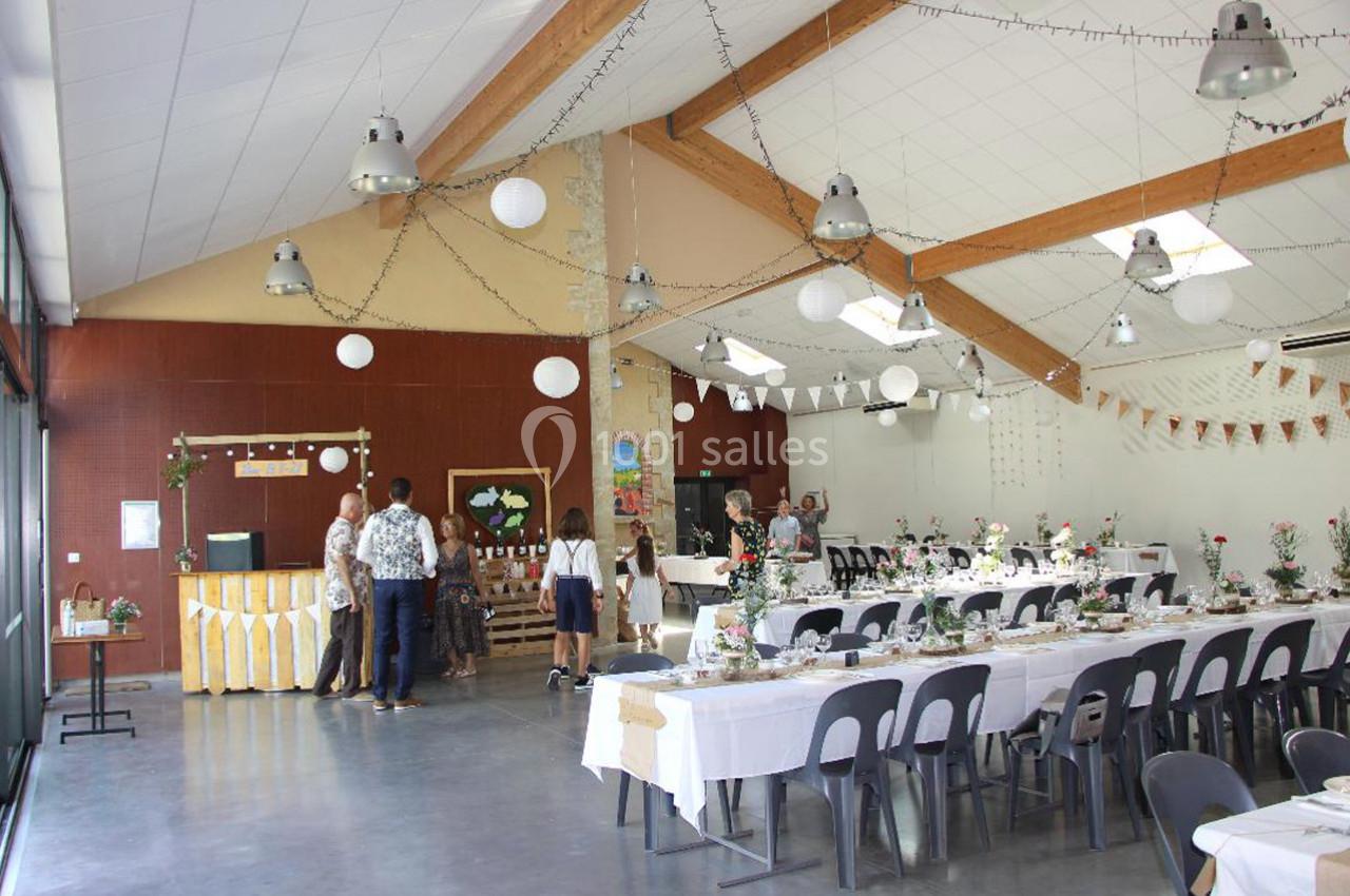 Salle décorée pour un événement, avec tables dressées, guirlandes lumineuses et invités discutant près d'un buffet.