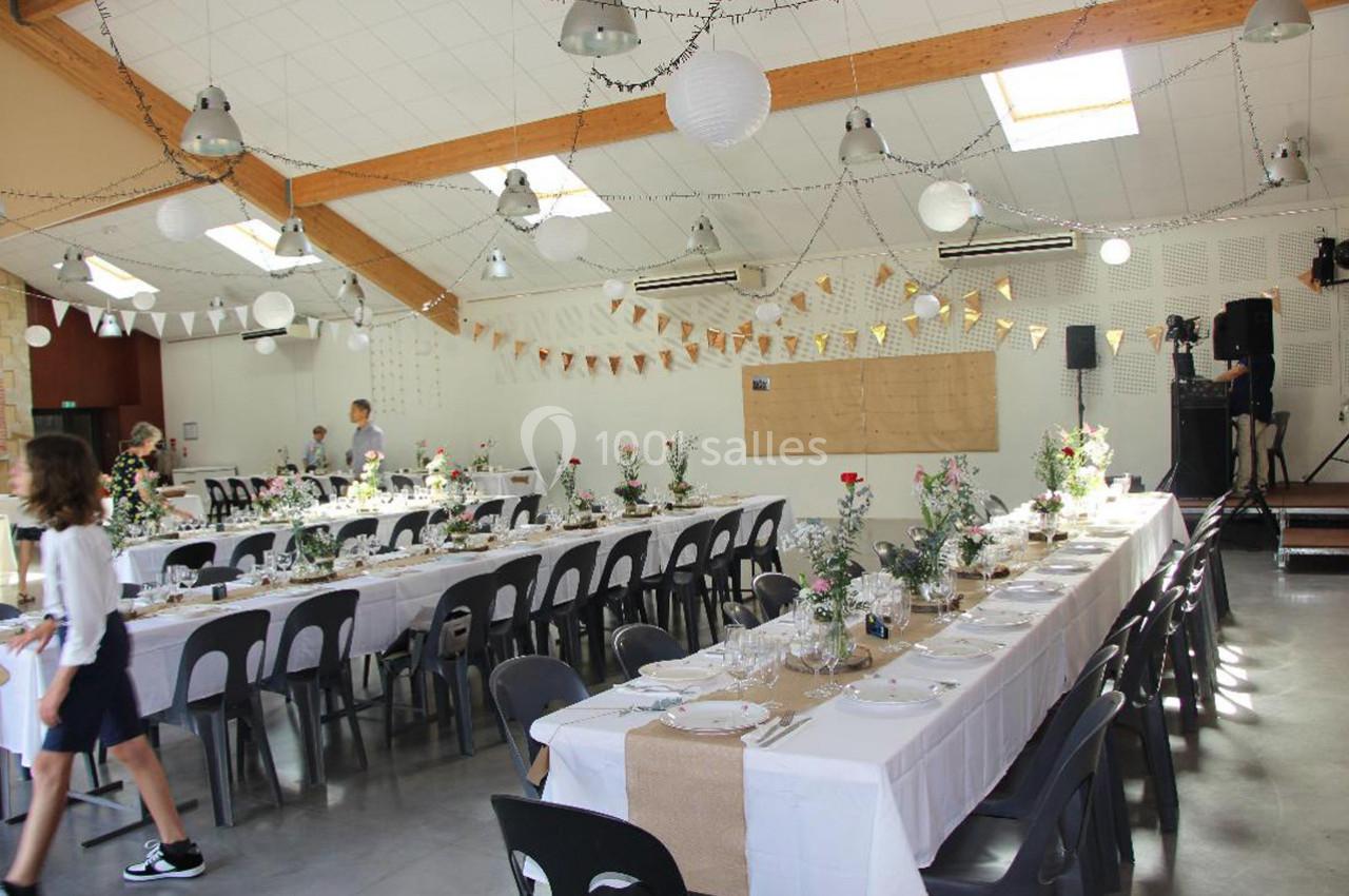 Salle décorée pour un événement avec de longues tables dressées, guirlandes lumineuses et fleurs en centre de table.