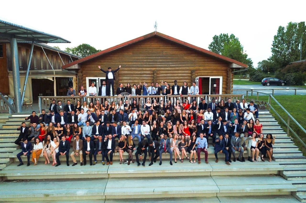 Groupe de personnes posant sur les marches devant un bâtiment en bois, dans un cadre extérieur verdoyant.