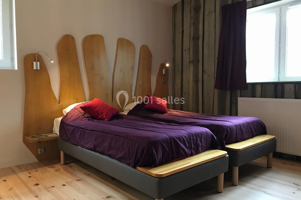 Deux lits simples avec draps violets, têtes de lit en bois sculpté, dans une chambre lumineuse avec parquet et mur en bois.