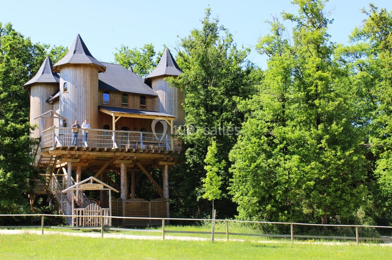 Cabane en bois sur pilotis avec des tours, entourée d'arbres et située dans un espace naturel verdoyant.