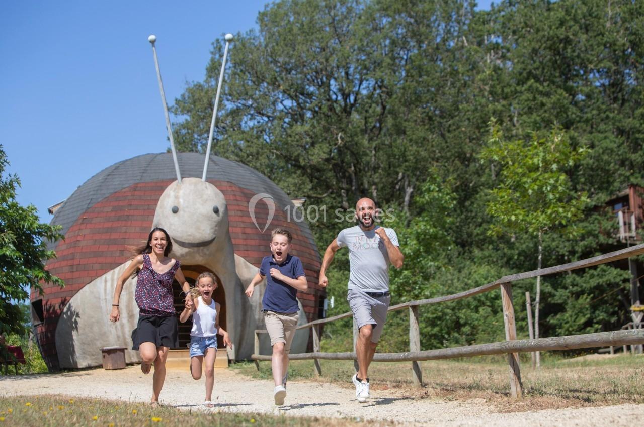 Une famille court sur un chemin en gravier devant une grande sculpture en forme d'escargot dans un cadre naturel.