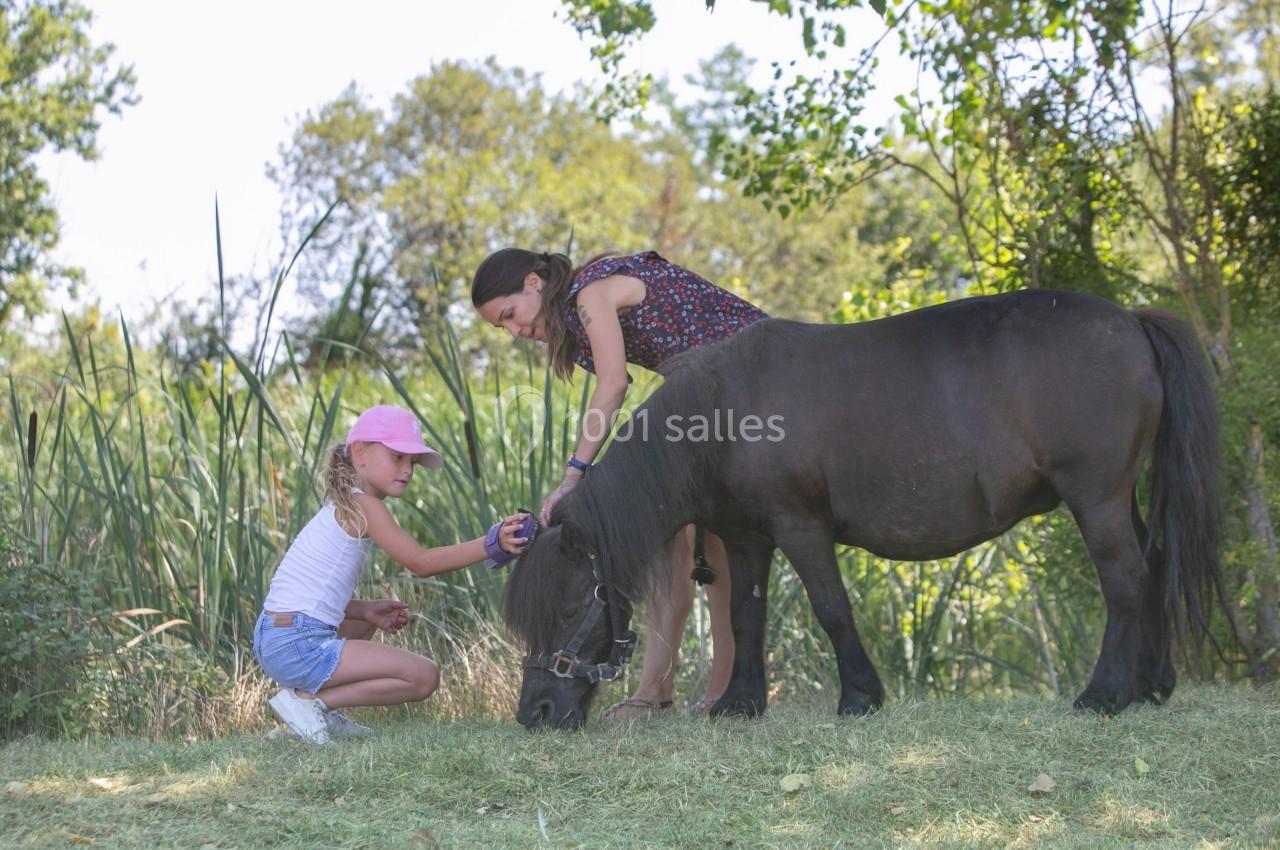 Une femme et une fillette caressent un poney noir broutant dans un espace herbeux entouré de végétation.