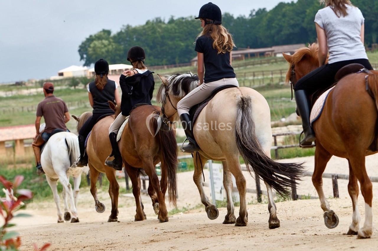 Groupe de cavaliers montant des chevaux sur un chemin en plein air, entouré de verdure et de bâtiments ruraux.