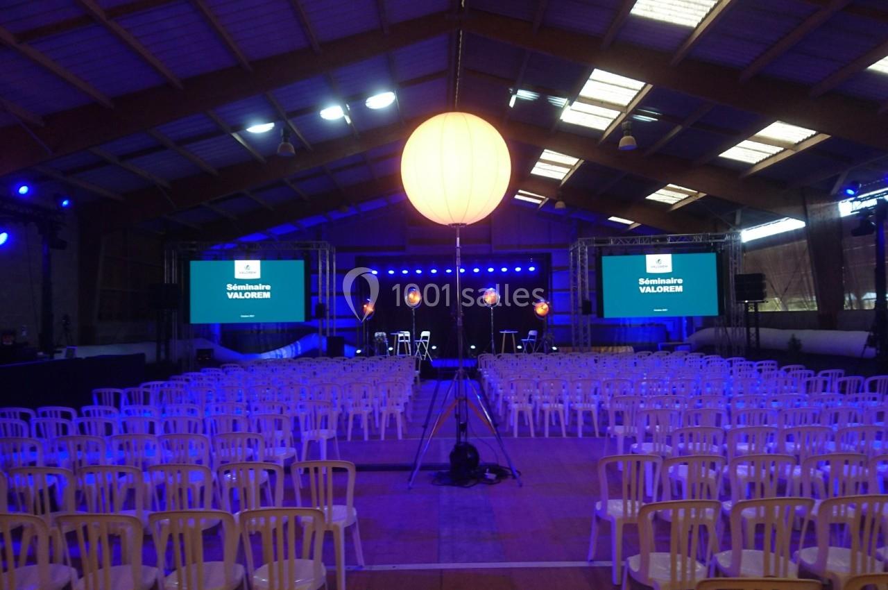 Salle de conférence avec des rangées de chaises blanches, scène éclairée en bleu et écrans affichant des présentations.