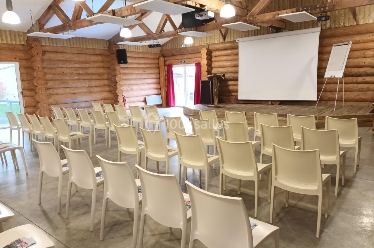 Salle de conférence en bois avec des rangées de chaises blanches, un écran de projection et un chevalet.