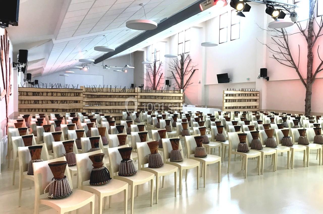 Salle lumineuse aménagée avec des rangées de chaises blanches, chacune équipée d'un djembé en bois.