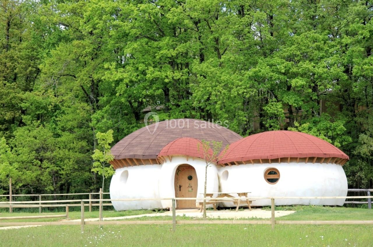 Maison en forme de champignon avec toit arrondi, entourée d'arbres et d'une clôture en bois, sur une pelouse.