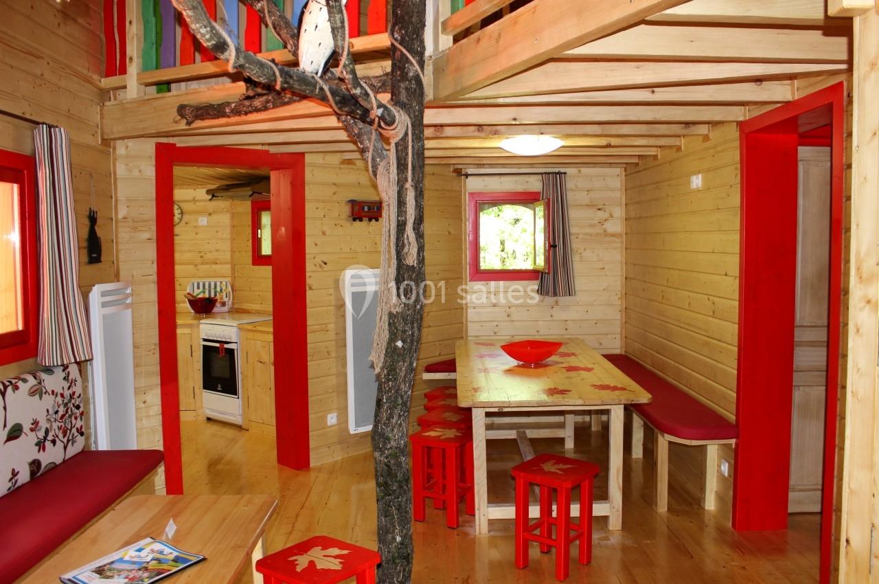 Intérieur d'une cabane en bois avec mobilier rouge, table à manger, cuisine ouverte et arbre traversant la pièce.
