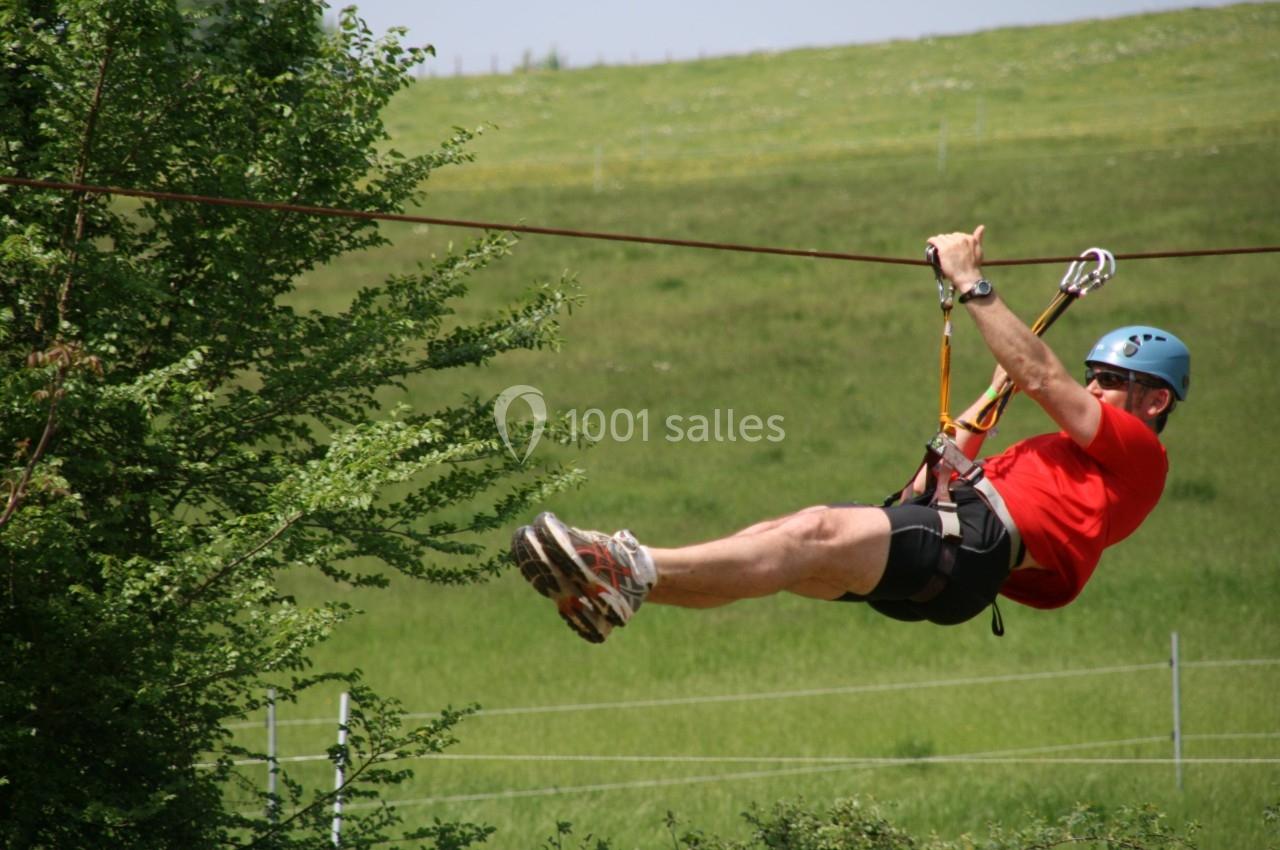 Un homme portant un casque et un harnais glisse sur une tyrolienne au-dessus d'un paysage verdoyant.