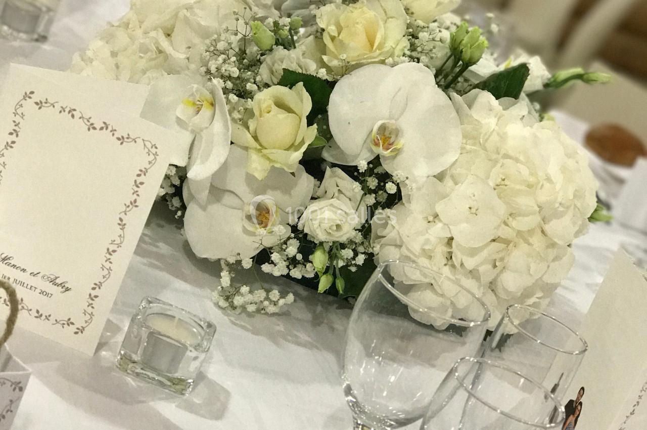 Centre de table floral blanc avec roses, orchidées et hortensias, entouré de verres et menus sur une nappe blanche.