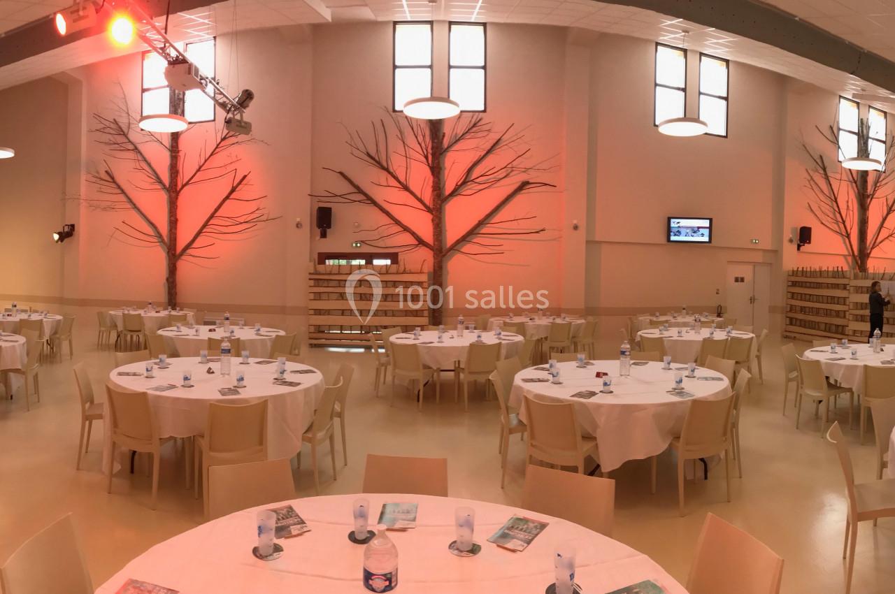 Salle de réception lumineuse avec tables rondes dressées, décorations murales en forme d'arbres et éclairage chaleureux.