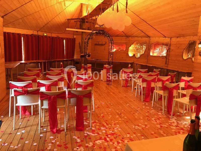 Salle en bois décorée pour une cérémonie, avec des chaises blanches ornées de nœuds rouges et des pétales au sol.