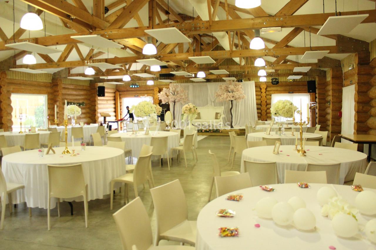 Salle de réception décorée avec des tables rondes, nappes blanches, fleurs et structure en bois apparente.
