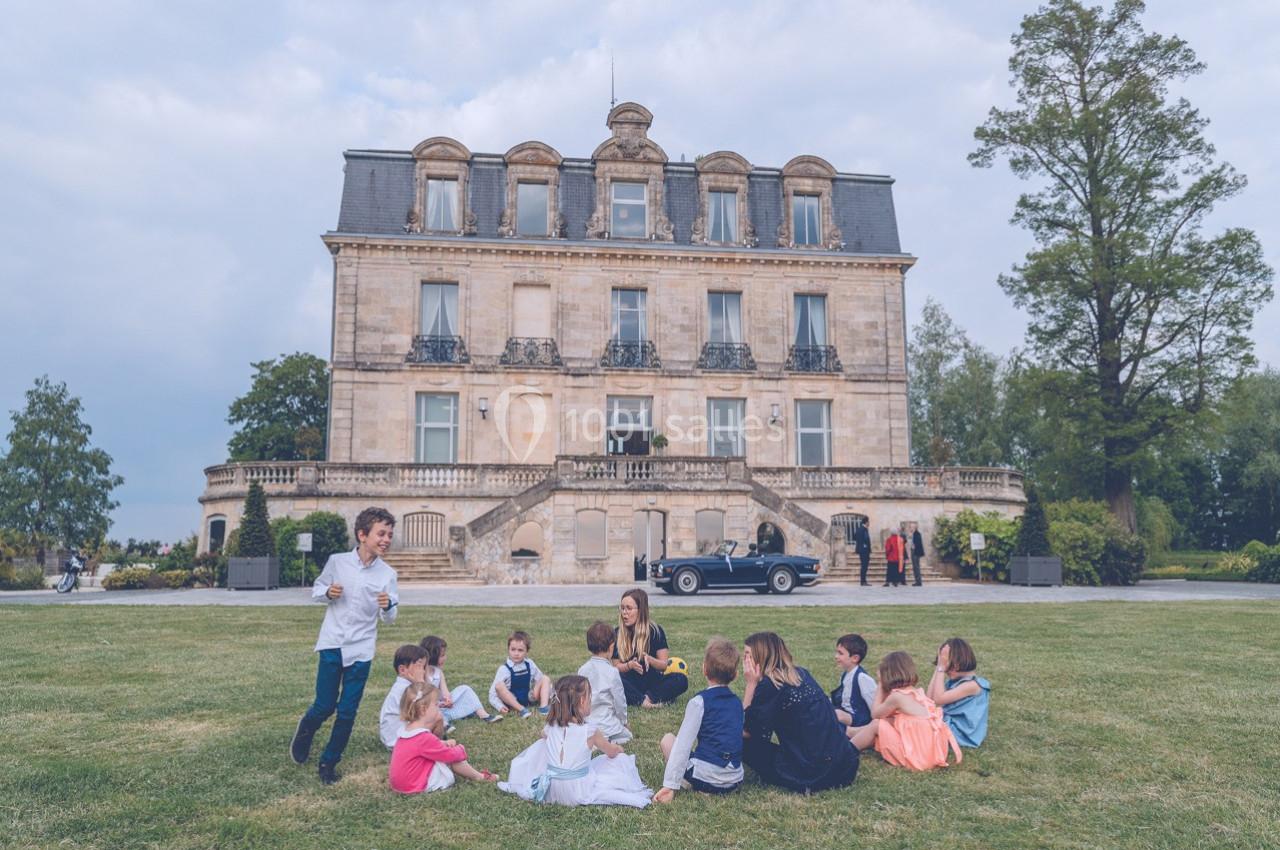 Un groupe d'enfants assis sur une pelouse devant un grand bâtiment ancien, avec des adultes à proximité.