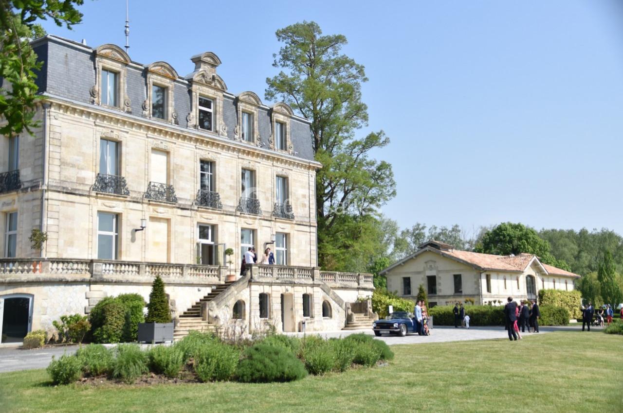 Façade d'un manoir en pierre avec un escalier, entouré de verdure et d'annexes sous un ciel dégagé.