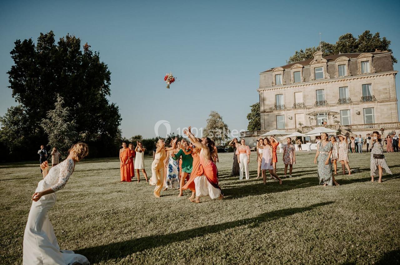 Un groupe de femmes tente d'attraper un bouquet lancé par une mariée dans un jardin devant un grand bâtiment.