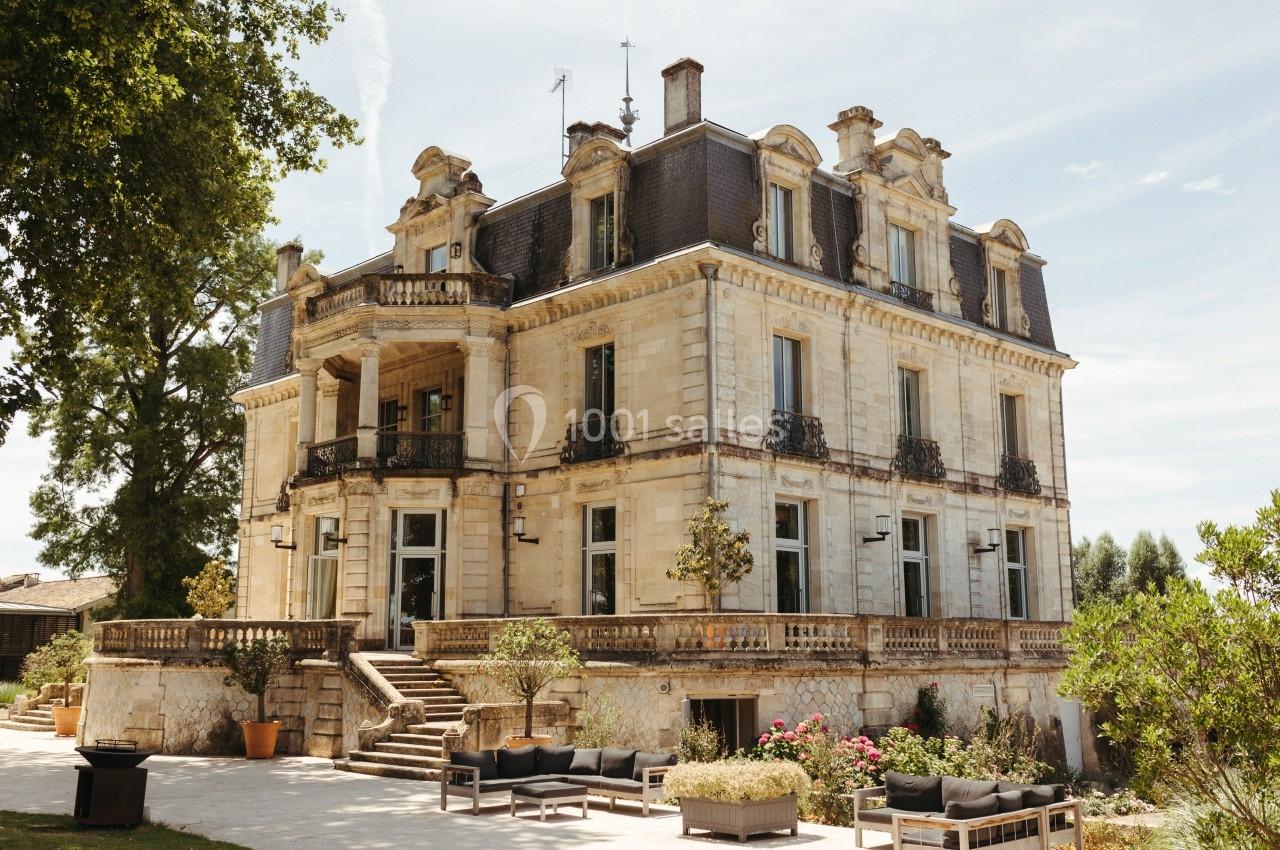 Façade d'un château en pierre avec balcons en fer forgé, entouré de végétation et de mobilier d'extérieur.