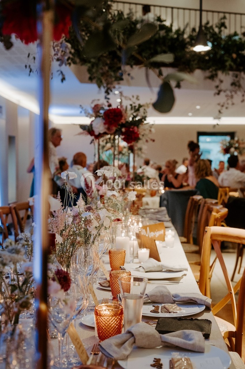 Table de réception décorée avec des bougies, fleurs et nappes blanches, entourée d'invités dans une salle éclairée.