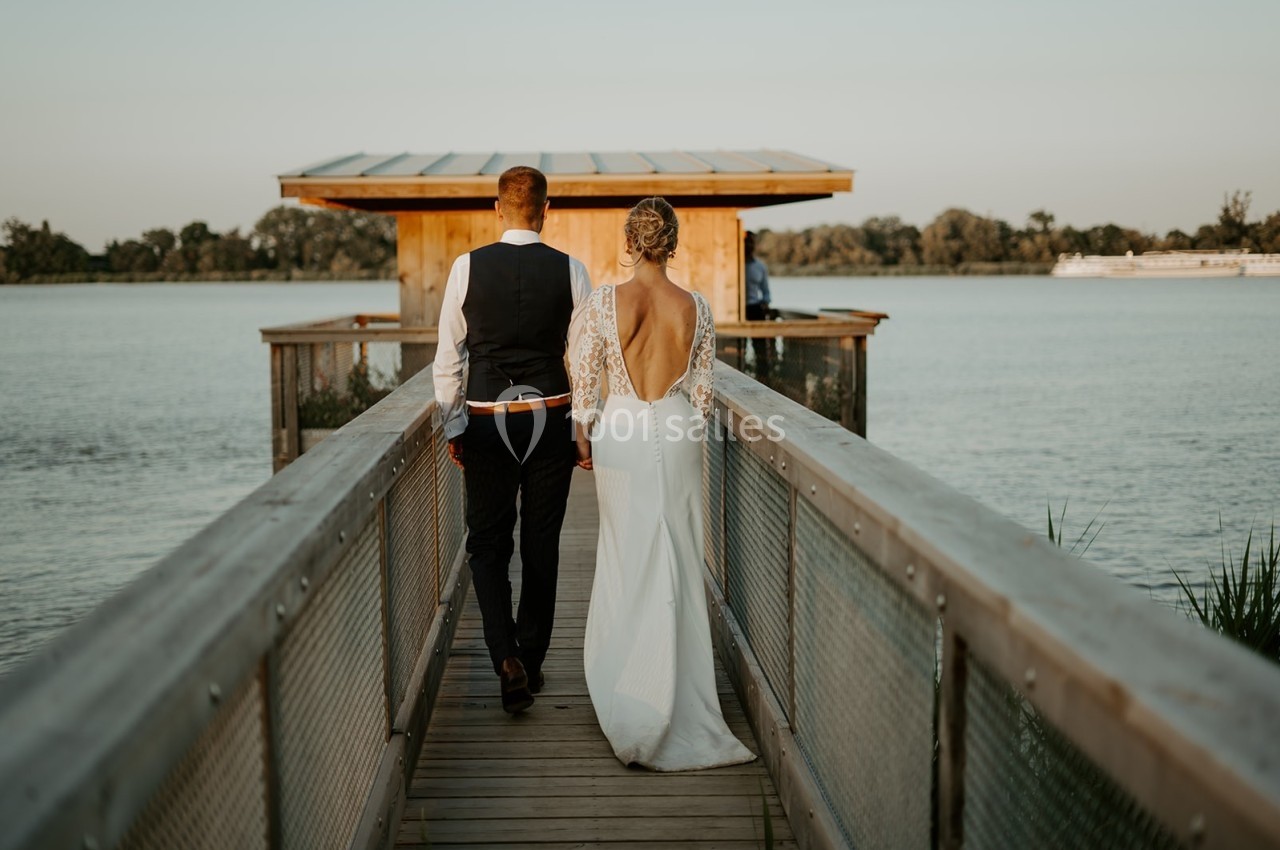 Un couple en tenue de mariage marche sur un ponton en bois menant à une cabane au bord d'un lac.