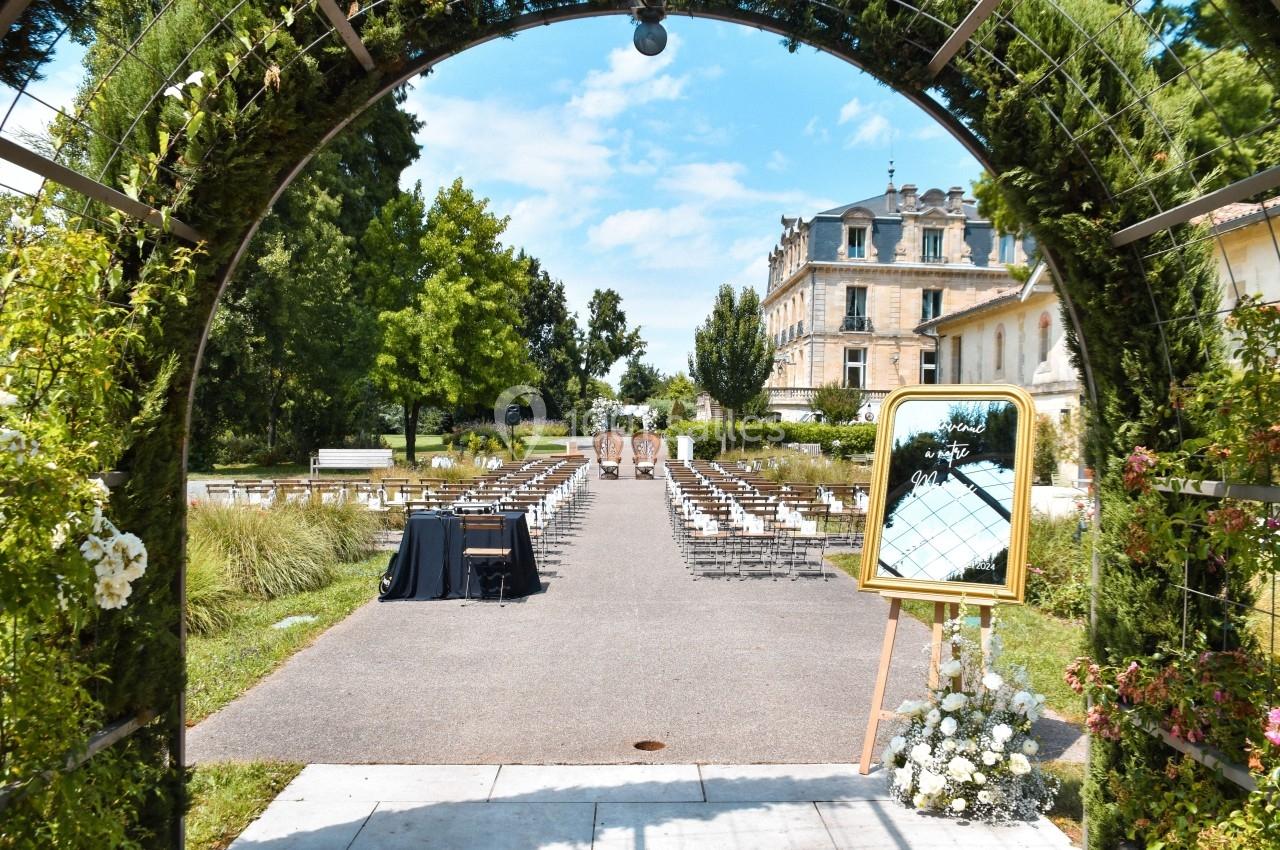 Allée extérieure aménagée pour une cérémonie, encadrée par une arche végétale, avec un château en arrière-plan.