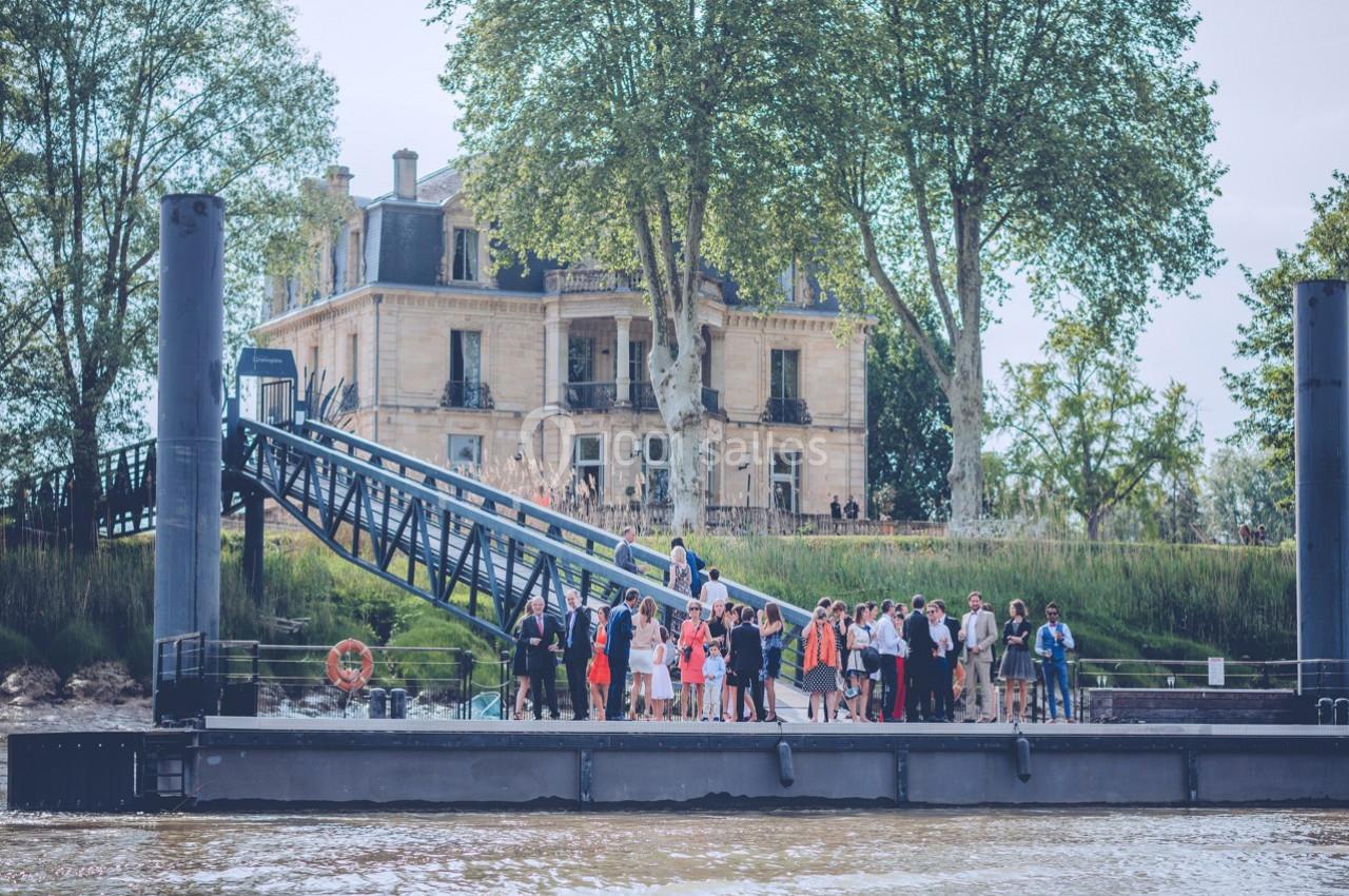 Groupe de personnes rassemblées sur un ponton au bord d'une rivière, avec un grand bâtiment en arrière-plan.
