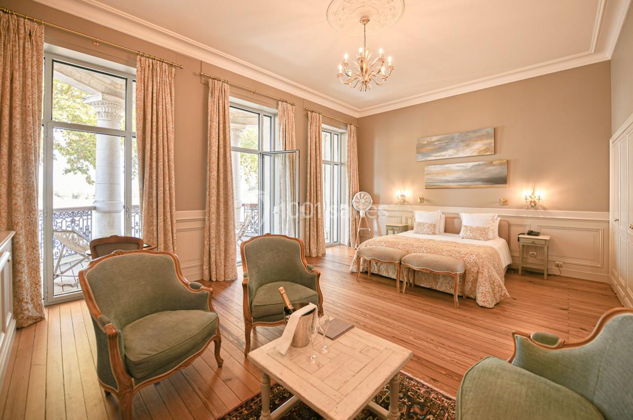 Chambre lumineuse avec lit double, fauteuils, table basse et grandes fenêtres donnant sur un balcon.