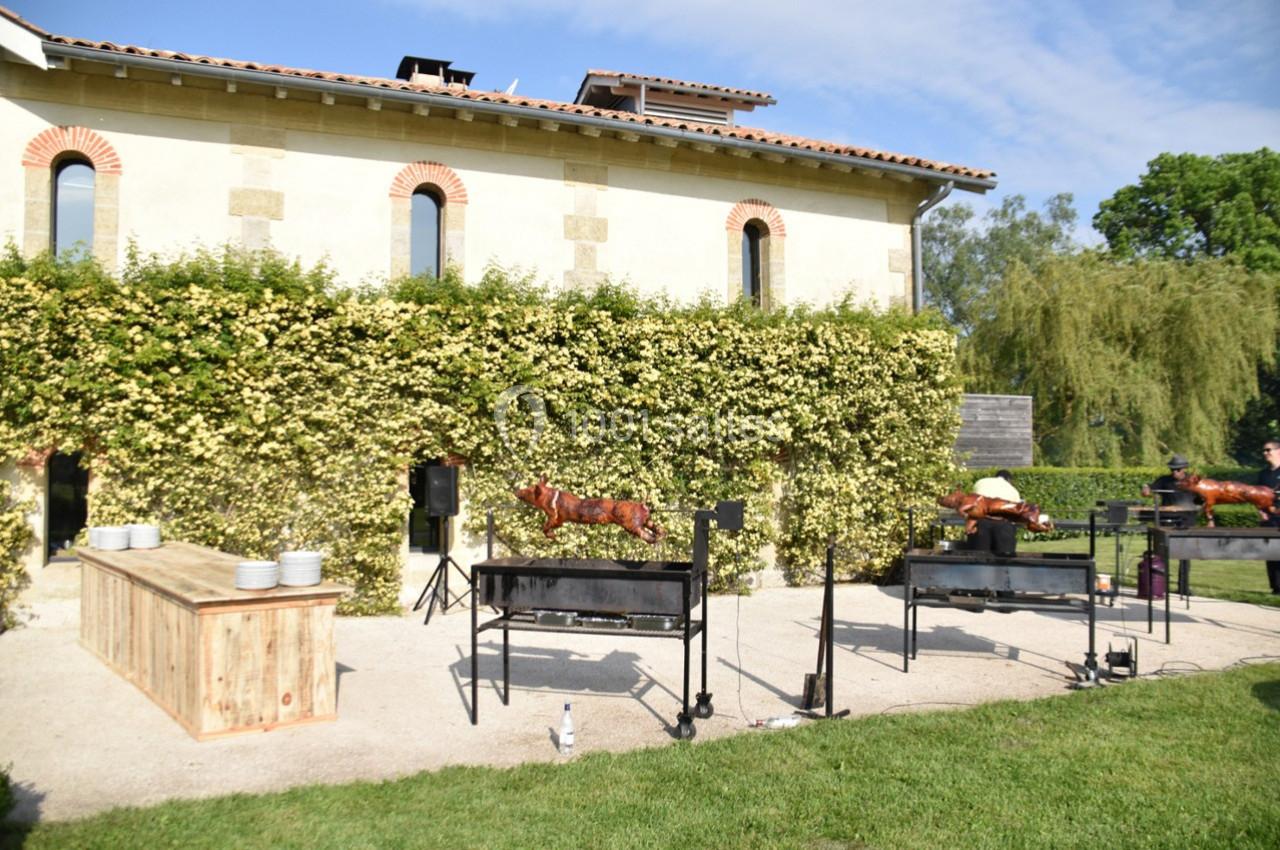 Des cochons de lait rôtissent sur des broches devant un bâtiment en pierre entouré de verdure.