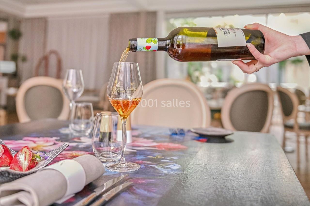 Une main verse du vin blanc dans un verre sur une table dressée avec une nappe fleurie dans un restaurant lumineux.