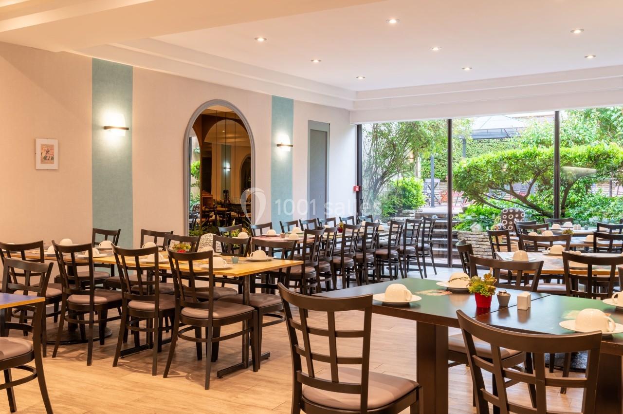 Salle lumineuse avec tables et chaises en bois, grandes baies vitrées donnant sur un jardin verdoyant.