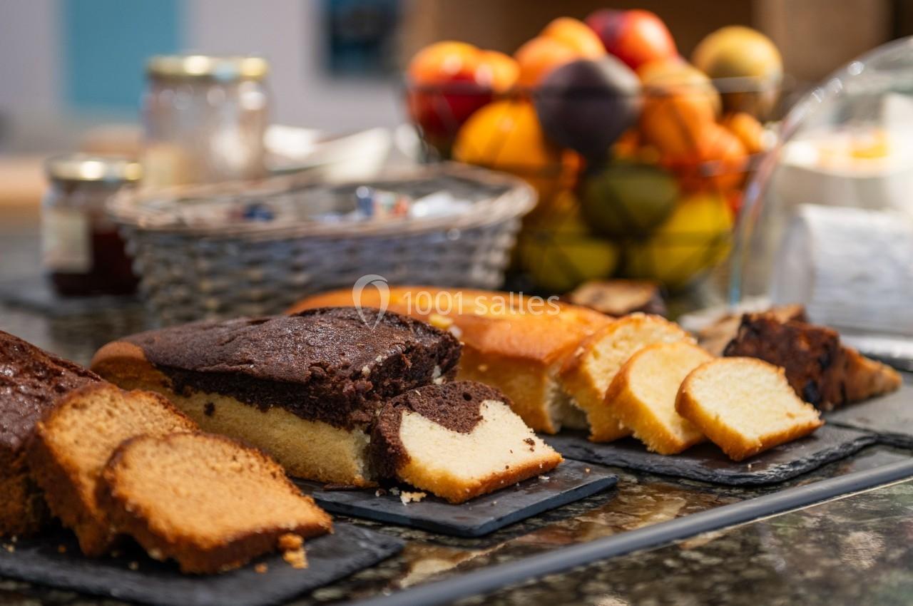 Assortiment de gâteaux tranchés disposés sur des ardoises, avec des fruits et des pots en arrière-plan.