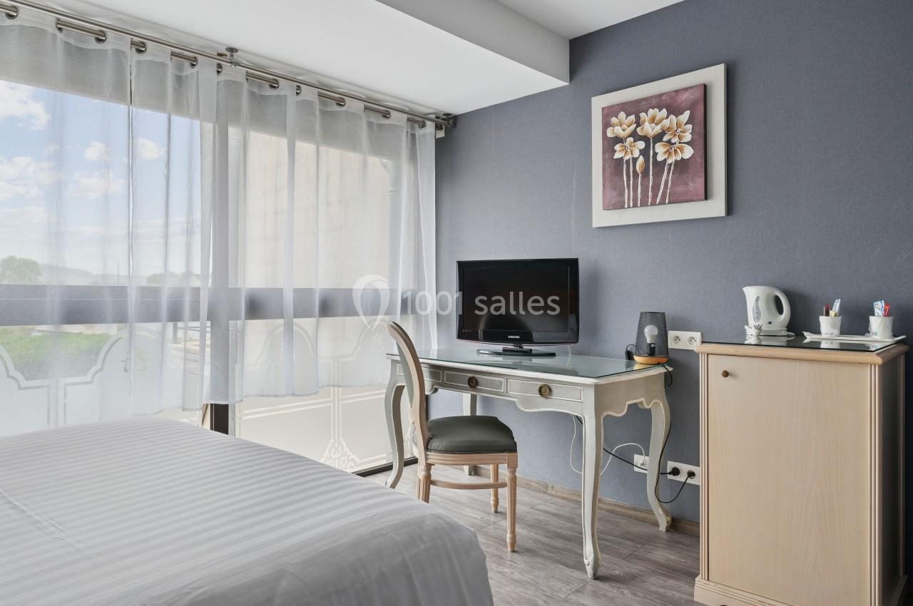 Chambre lumineuse avec bureau, télévision, bouilloire et grande fenêtre avec rideaux translucides.